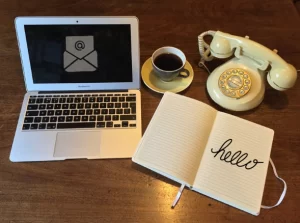 A phone, a notebook with the text 'hello', and a laptop screen with email icon, all shown to suggest contacting